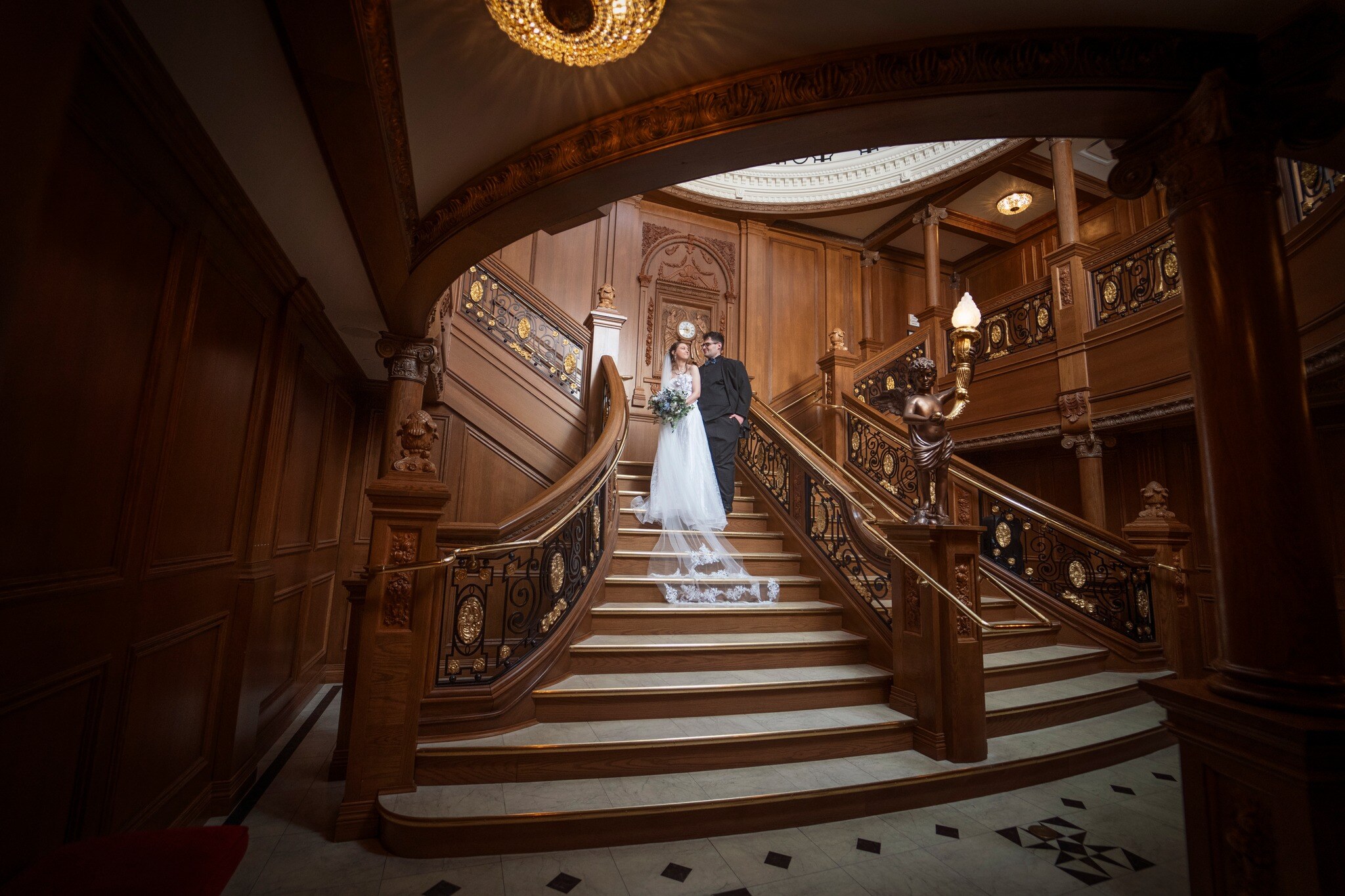 A bride and groom dressed in suit and wedding dress stand on the Grand Staircase of the Titanic Museum Attraction after their wedding ceremony.