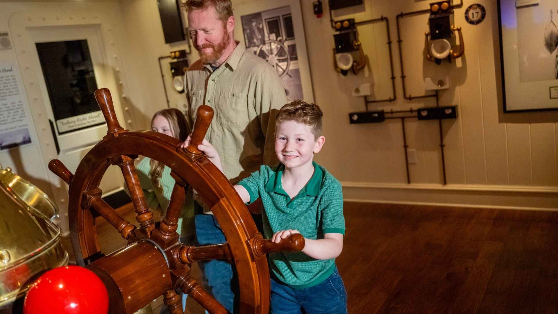 Young boy and his father are on the Captain's Bridge, turning the wheel while looking out at the starry night in this interactive gallery.