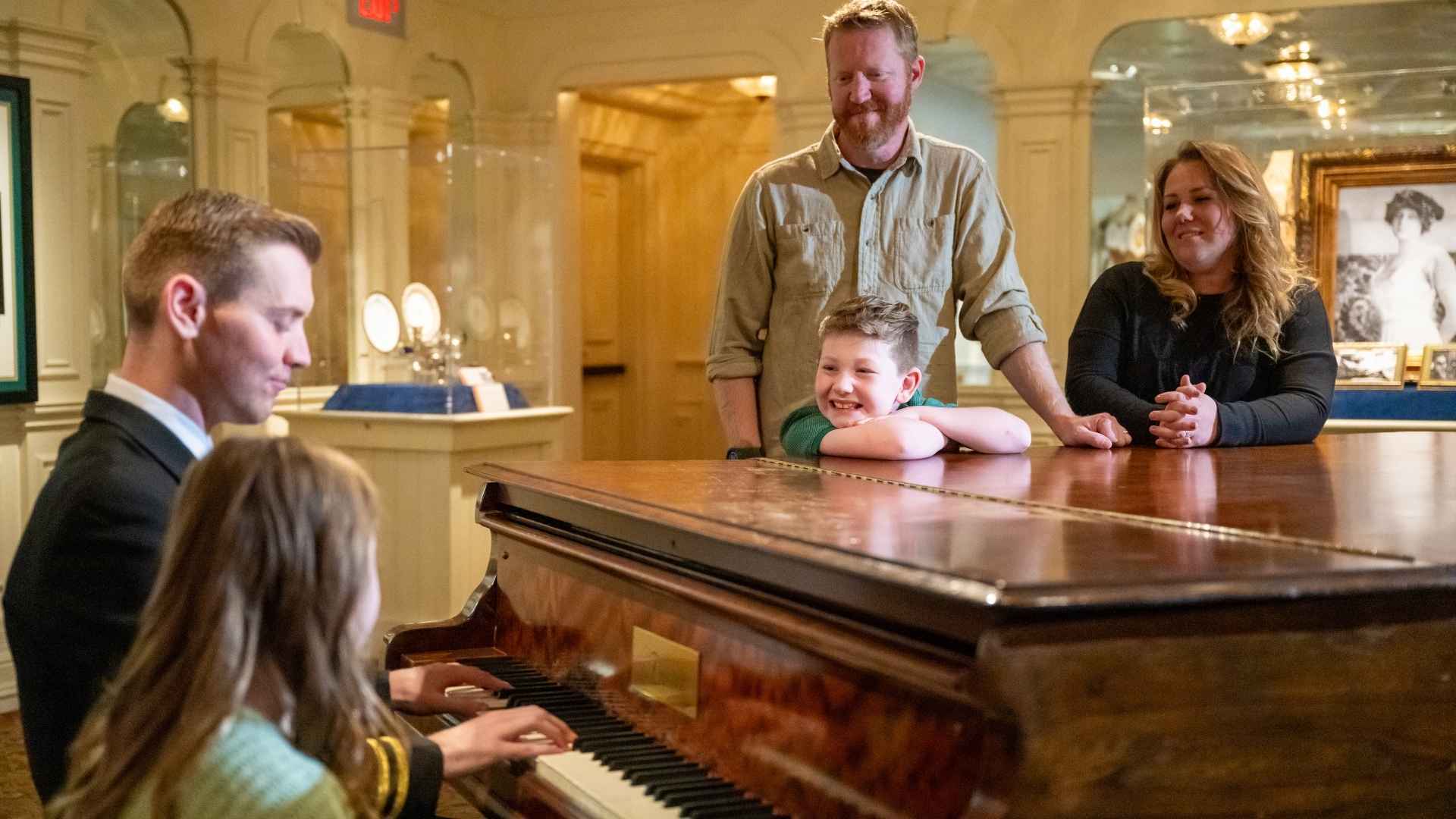 Family gathered around a piano while crew member plays a period song.